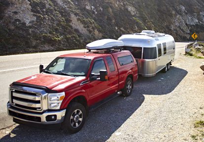 Auto Plus Insurance Group LLC | Red truck parked on the side of a road near a mountain with a silver RV attached to the back and a luggage rack on top